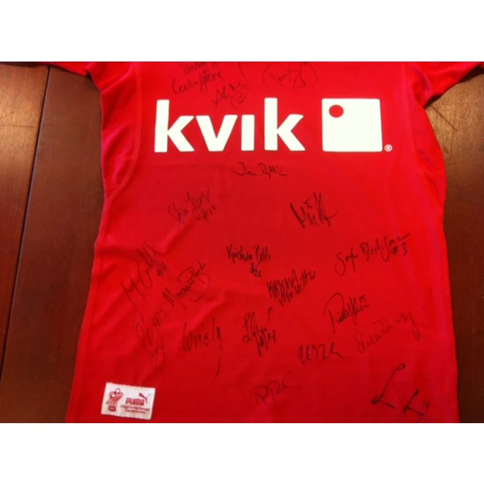 En rød skjorte med det hvide kvik-logo og flere sorte signaturer skrevet hen over fronten. Skjorten er lagt fladt på en træoverflade.